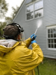 Person in a yellow rain jacket using a pressure washer to clean the exterior of a house.